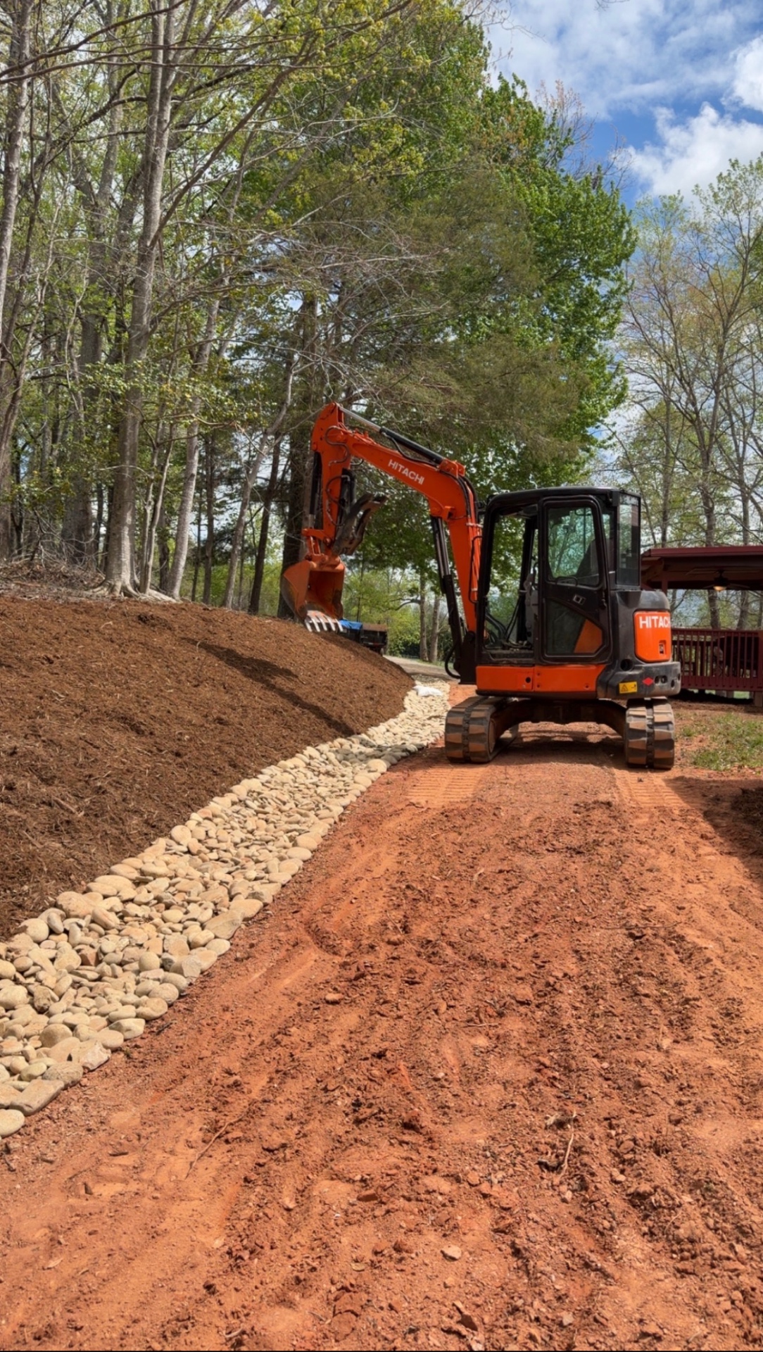 Orange Hitachi excavator beside a freshly installed stone drainage channel on a red clay driveway